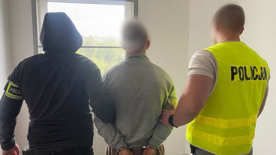 Dwóch policjantów prowadzi zatrzymanego mężczyznę w kajdankach, twarze zamazane.