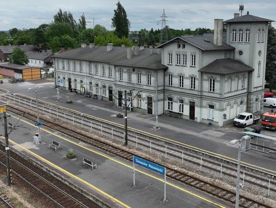Panorama stacji kolejowej Tomaszów Mazowiecki z budynkiem dworca, peronami i torami; widoczne oznaczenia peronów i pojazdy serwisowe podczas modernizacji.