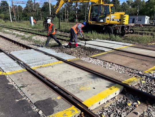 Zbliżenie na torowisko w Tomaszowie Mazowieckim; robotnicy montują płyty między szynami, w tle koparka, żółte oznaczenia krawędzi peronu.
