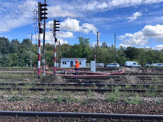 Szerokie ujęcie torów stacji Tomaszów Mazowiecki z pracownikiem wykonującym prace kablowe obok semaforów; widoczny teren budowy i niebo z chmurami.
