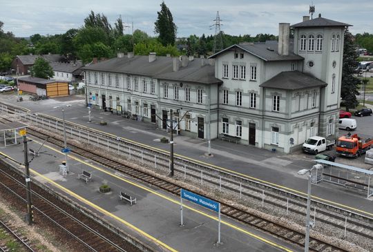 Panorama stacji kolejowej Tomaszów Mazowiecki z budynkiem dworca, peronami i torami; widoczne oznaczenia peronów i pojazdy serwisowe podczas modernizacji.