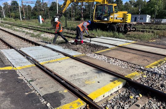 Zbliżenie na torowisko w Tomaszowie Mazowieckim; robotnicy montują płyty między szynami, w tle koparka, żółte oznaczenia krawędzi peronu.