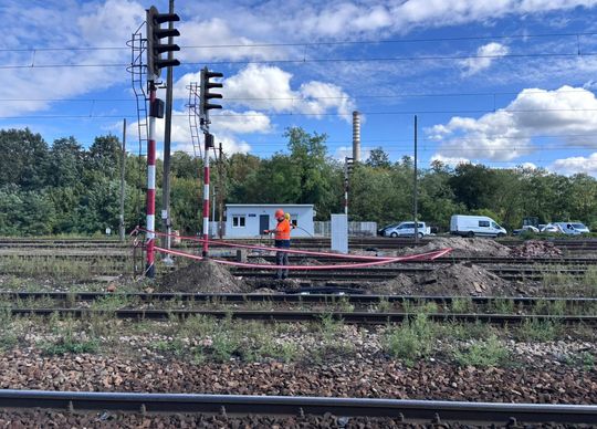 Szerokie ujęcie torów stacji Tomaszów Mazowiecki z pracownikiem wykonującym prace kablowe obok semaforów; widoczny teren budowy i niebo z chmurami.