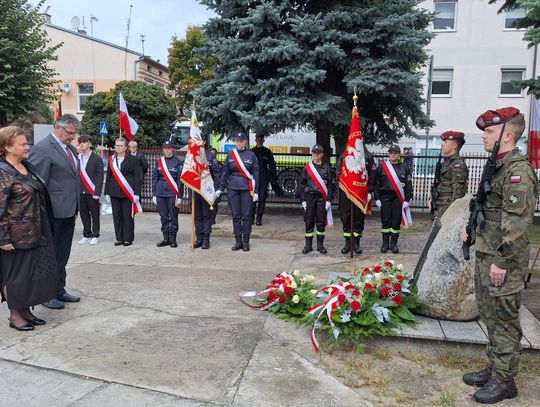 Delegacja składa wieniec przy kamiennym obelisku, asysta wojskowa w szyku