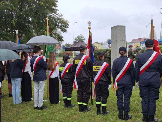 Uczestnicy z pocztami i klasy pożarnicze stoją przy pomniku pod parasolami
