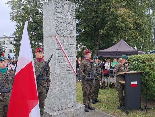 Żołnierze w umundurowaniu z bronią stoją przy pomniku, obok mównica i polskie flagi