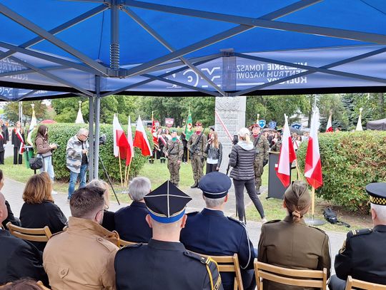 Uczestnicy pod namiotem obserwują uroczystość, widoczne flagi państwowe i obelisk