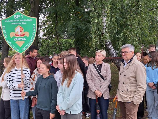 Uczniowie SP1 z tablicą szkoły stoją pod drzewami podczas uroczystości