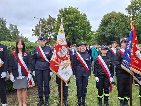 Szereg uczniów w oliwkowych mundurach obok biało-czerwonego wieńca