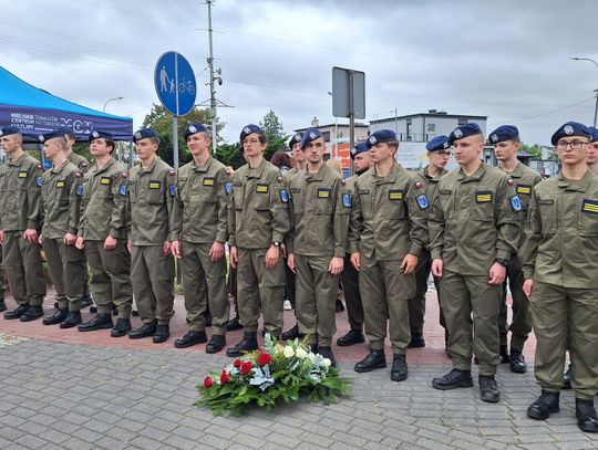 Szereg uczniów w oliwkowych mundurach obok biało-czerwonego wieńca