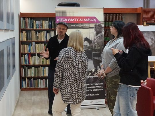 Zwiedzający rozmawiają przy ekspozycji w bibliotece; w tle plakat projektu „Niech fakty zatańczą” i regały z książkami.