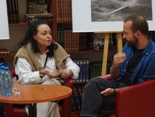 Dyskusja dwojga uczestników na tle czarno-białych fotografii w czytelni Miejskiej Biblioteki Publicznej w Tomaszowie Mazowieckim.