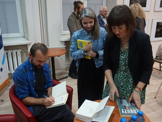 Uczestnicy czekają na podpis w książkach; widoczny roll-up Miejskiej Biblioteki Publicznej i ekspozycja czarno-białych fotografii.