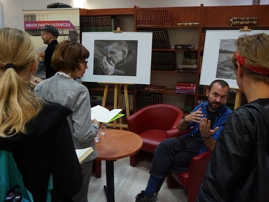 Autor rozmawia z uczestnikami obok plansz z czarno-białymi fotografiami w przestrzeni Miejskiej Biblioteki Publicznej.