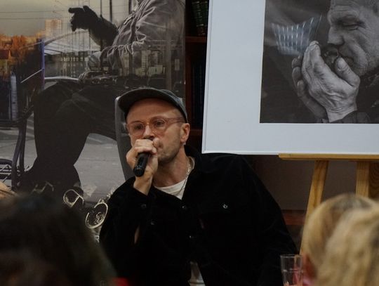 Mówca z mikrofonem na tle plakatu projektu i fotografii przedstawiającej zamyślonego mężczyznę.