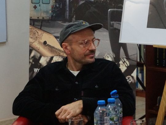 Mężczyzna w czapce siedzi i słucha; na pierwszym planie butelki wody, w tle fotografia dokumentalna.