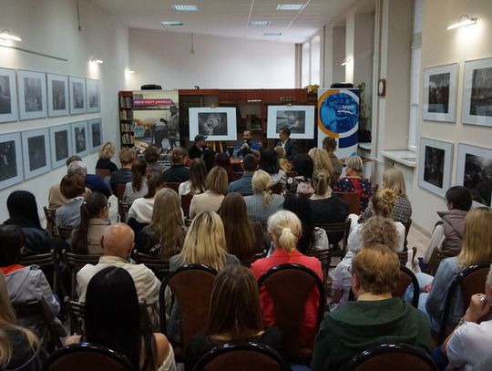 Szeroki kadr biblioteki; publiczność, rząd czarno-białych fotografii na ścianach i panel dyskusyjny pośrodku.