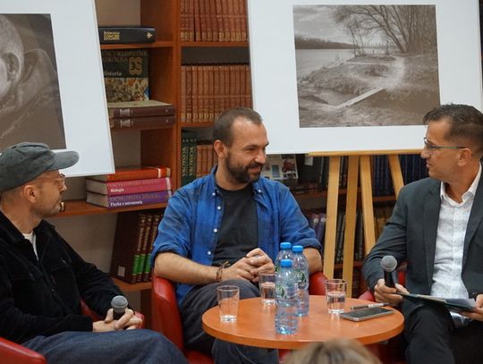 Trzech panelistów przy stoliku z mikrofonami; za nimi czarno-białe zdjęcia na sztalugach i regały z książkami