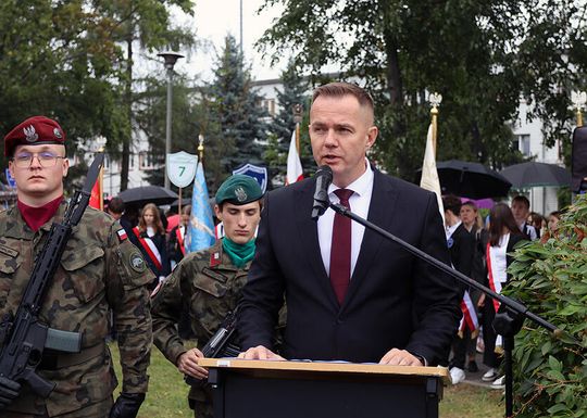 Wystąpienie przy mównicy, w tle żołnierze i poczty sztandarowe