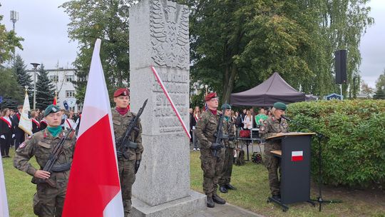 Żołnierze w umundurowaniu z bronią stoją przy pomniku, obok mównica i polskie flagi
