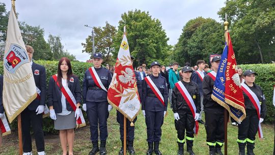 Szereg uczniów w oliwkowych mundurach obok biało-czerwonego wieńca