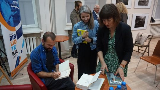 Uczestnicy czekają na podpis w książkach; widoczny roll-up Miejskiej Biblioteki Publicznej i ekspozycja czarno-białych fotografii.