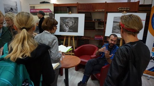 Autor rozmawia z uczestnikami obok plansz z czarno-białymi fotografiami w przestrzeni Miejskiej Biblioteki Publicznej.