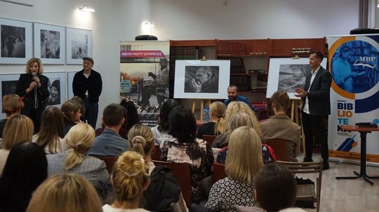 Prelegenci przed publicznością; czarno-białe fotografie na sztalugach, banery MBP i „Niech fakty zatańczą” w czytelni.