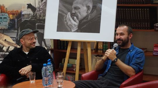 Dwóch autorów, w tle czarno-białe zdjęcie i regały biblioteki.