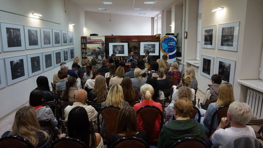 Szeroki kadr biblioteki; publiczność, rząd czarno-białych fotografii na ścianach i panel dyskusyjny pośrodku.