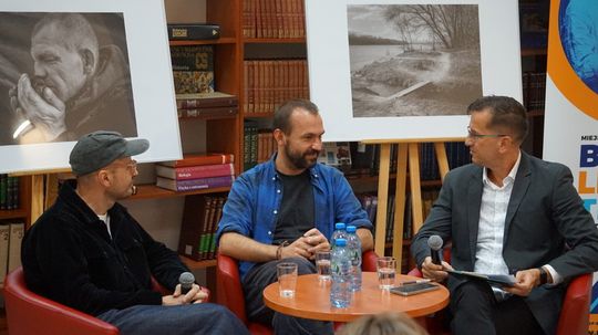 Trzech panelistów przy stoliku z mikrofonami; za nimi czarno-białe zdjęcia na sztalugach i regały z książkami