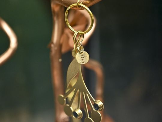 Zbliżenie na pozłacany brelok z elementami ceramicznymi zawieszony na metalowej gałęzi.