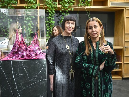 Dwie uczestniczki stoją obok gabloty z różowo-fuksjową instalacją i pozłacanymi brelokami.