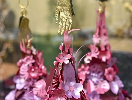 Pozłacana zawieszka w kształcie dłoni nad papierową instalacją z różowo-fioletowych kwiatów.