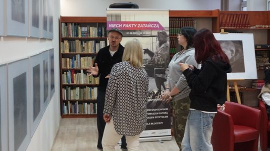 Zwiedzający rozmawiają przy ekspozycji w bibliotece; w tle plakat projektu „Niech fakty zatańczą” i regały z książkami.