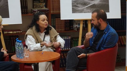 Dyskusja dwojga uczestników na tle czarno-białych fotografii w czytelni Miejskiej Biblioteki Publicznej w Tomaszowie Mazowieckim.