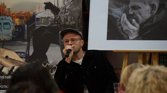 Mówca z mikrofonem na tle plakatu projektu i fotografii przedstawiającej zamyślonego mężczyznę.