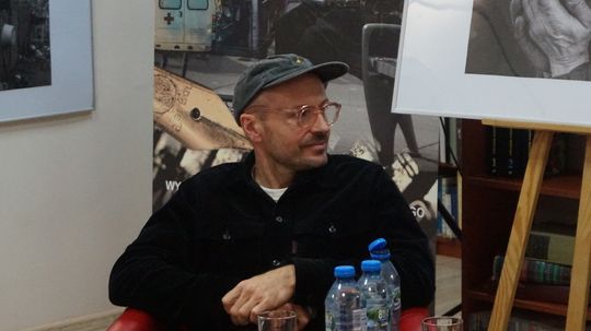 Mężczyzna w czapce siedzi i słucha; na pierwszym planie butelki wody, w tle fotografia dokumentalna.