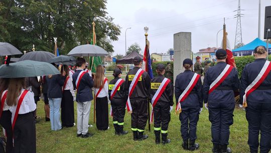 Poczty sztandarowe przy pomniku Sybiraków w Tomaszowie Uczestnicy z pocztami i klasy pożarnicze stoją przy pomniku pod parasolami