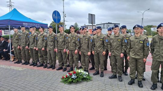 Klasy mundurowe w szyku przy złożonym wieńcu Szereg uczniów w oliwkowych mundurach obok biało-czerwonego wieńca