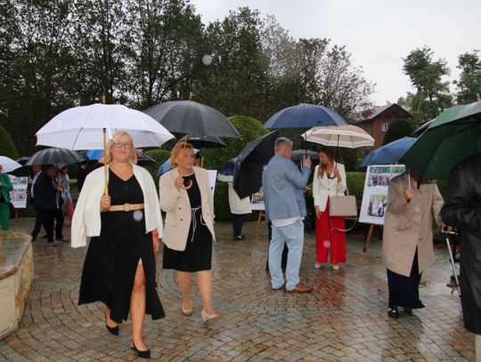 Uczestnicy z parasolami idą alejką parku, oglądając plenerową ekspozycję o dożynkach.