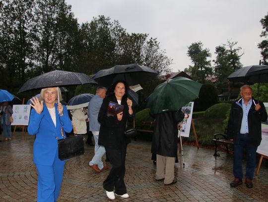 Kobieta macha do aparatu; wokół goście z parasolami i plansze wystawy w parku.