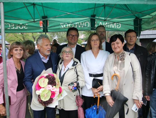 Grupa uczestników pozuje pod zielonym namiotem, w centrum bukiet kwiatów.