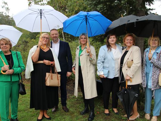 Siedem osób w eleganckich strojach stoi na trawie pod parasolami, pozując do zdjęcia podczas deszczowej uroczystości plenerowej.