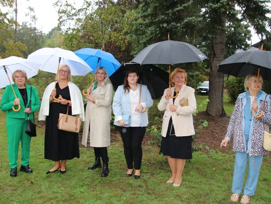 Siedem osób ustawionych w rzędzie z parasolami na tle drzew; pamiątkowe zdjęcie wykonane w deszczu.