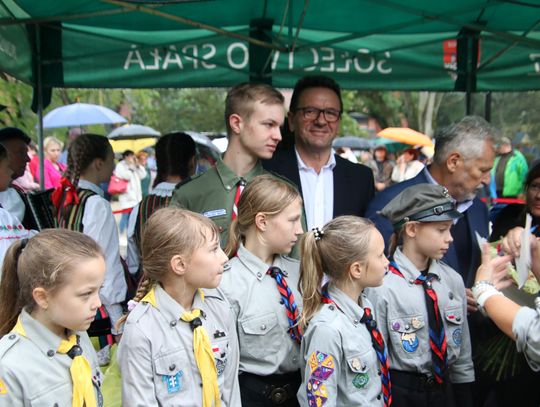 Dzieci w mundurach harcerskich z chustami stoją pod namiotem obok dorosłych gości podczas plenerowej uroczystości.