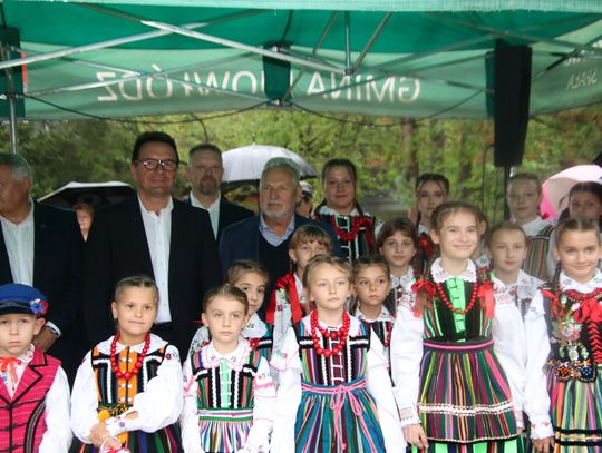 Grupa dzieci w kolorowych strojach ludowych stoi frontem do aparatu, za nimi dorośli pod namiotem.