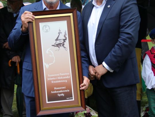 Dwóch mężczyzn trzyma dużą, oprawioną tablicę z napisem „Honorowi Ambasadorowie Spały” podczas ceremonii.