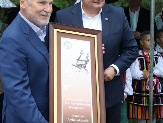 Dwie osoby pokazują do kamery oprawiony dyplom z napisem „Honorowi Ambasadorowie Spały”; w tle uczestnicy w strojach ludowych.