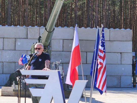 Wystąpienie na tle stanowiska artyleryjskiego Mówca przy mównicy z logo, obok flagi Polski i USA; za nim widać lufę haubicy i ścianę ochronną.